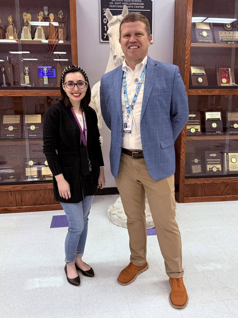 Two adults stand side by side in a school hallway in front of a glass trophy case filled with awards. One person, shorter, wears glasses, a black sweater, jeans, and a lanyard, smiling at the camera. The other person, taller, wears a blue blazer, white shirt, khaki pants, brown shoes, and a staff lanyard, also smiling. A white horse statue is positioned behind them, and school trophies and plaques are visible in the background.