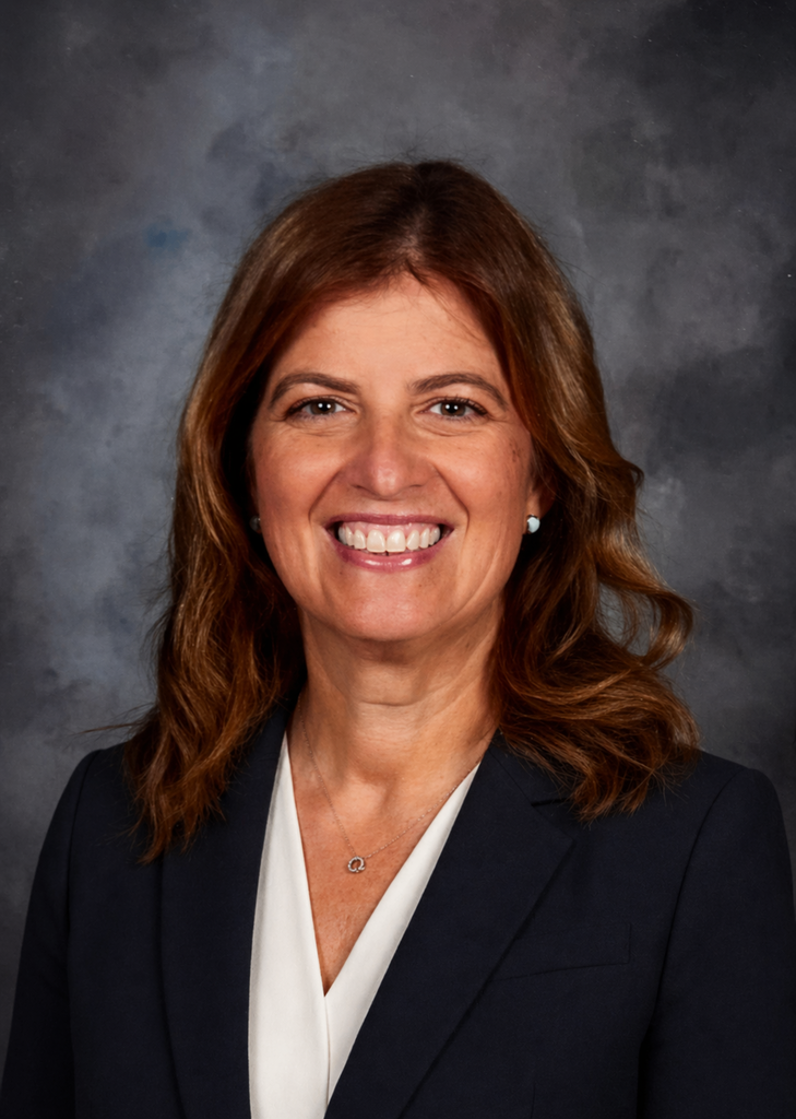 Professional head-and-shoulders portrait of Dr. Christie Dixon smiling at the camera. She has shoulder-length medium brown hair styled in loose waves and is wearing a navy blazer over a white blouse with small stud earrings and a delicate silver necklace. The portrait is set against a dark gray mottled studio background.