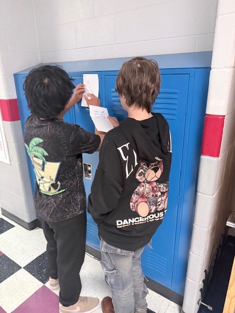 two students working on their kindness cards standing at a locker