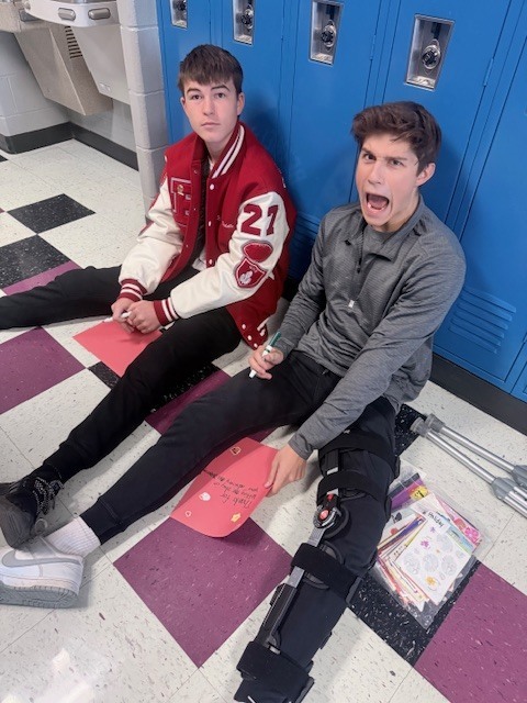 two male students sitting in the hall, one making a silly face