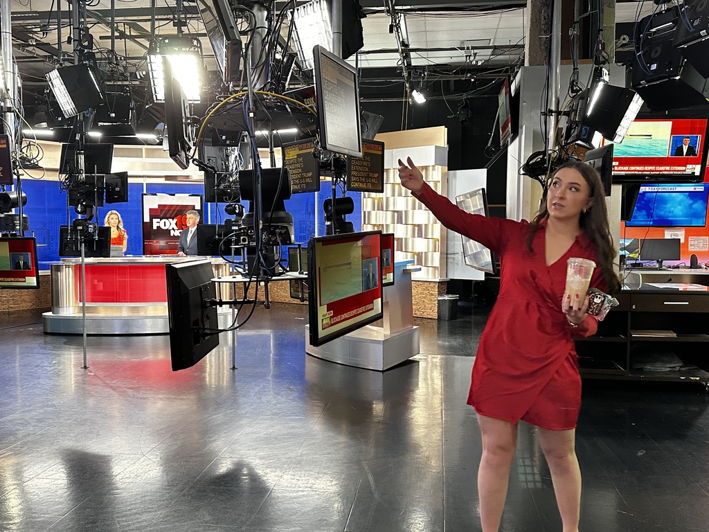 Bethel-Tate Content Creator students tour fox 19 news studio. As shown, gemale with one hand stretched out toward a monitor with studio in the background.