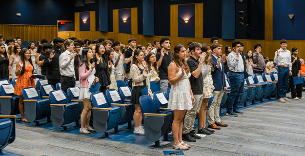 Students standing in auditorium, with one hand raised.