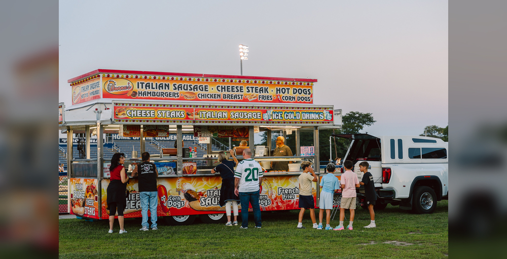 People gather at a food truck serving various items in a grassy area during an