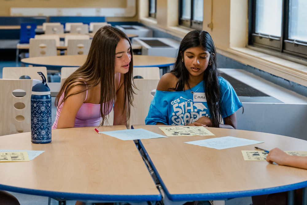 Two young girls collaborate at a classroom table, discussing papers and notes, with a water bottle nearby, in a bright learning environment.