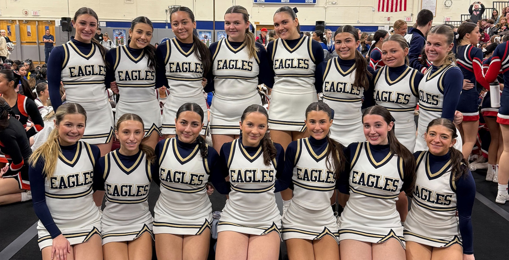 A group of cheerleaders dressed in matching white uniforms with Eagles logo stand together, smiling confidently in a gym setting.