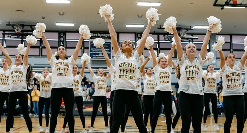Cheerleaders in matching "Bethpage Kickline" shirts energetically raise white pom-poms in a gym, conveying excitement and team spirit.