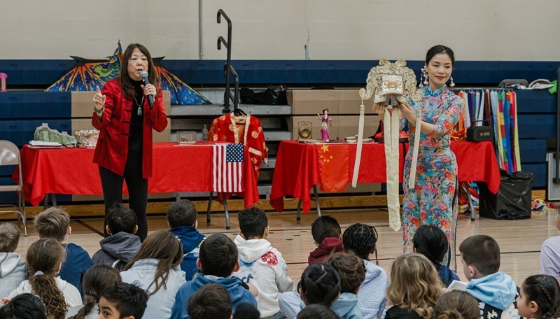 Two women present Chinese cultural items to seated children. One speaks into a microphone; the other holds a decorative headdress. Tables with flags, red cloths, and various objects are in the background. The mood is educational and engaging.