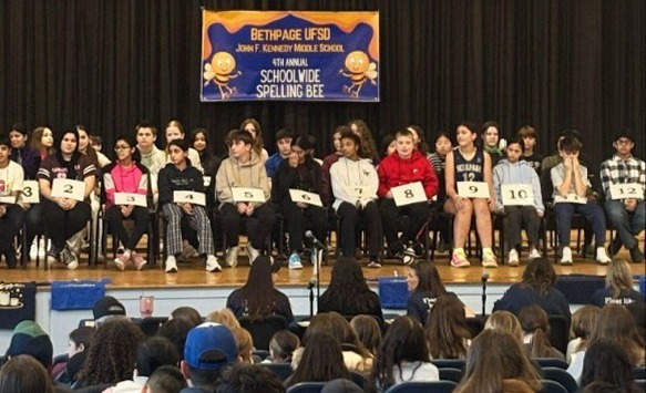 A group of students sit on a stage for a spelling bee, each with a numbered card. A banner overhead reads "4th Annual Schoolwide Spelling Bee." The audience watches attentively.
