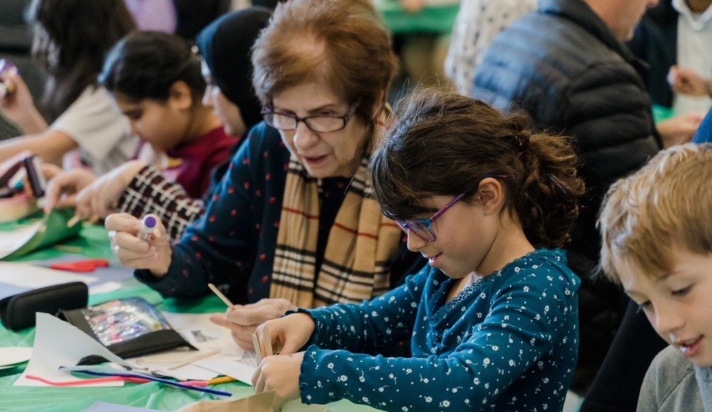 older woman and young girl engage in arts and crafts at a crowded table, fostering creativity and connection. A lively community atmosphere prevails.