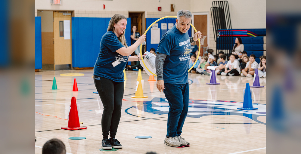Two people interact with a hula hoop in a gym while students watch.