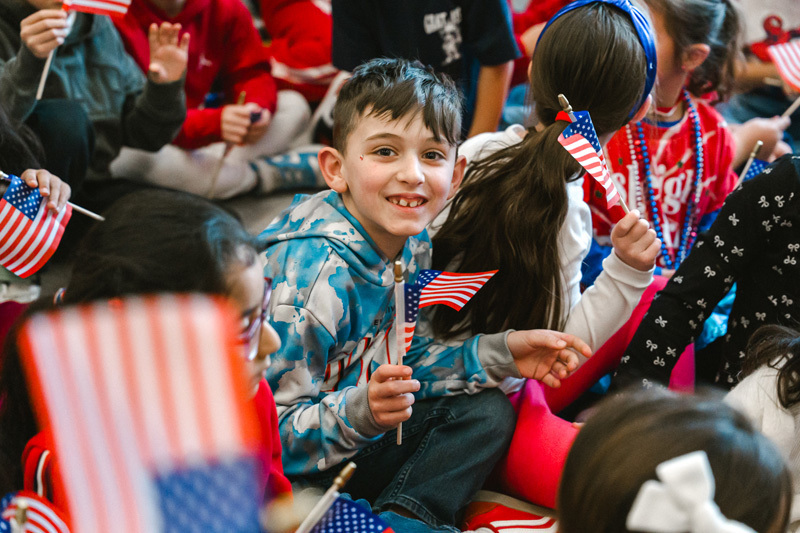 A diverse group of children sit closely together, some waving small American flags. A boy in a red shirt is smiling brightly, creating a joyful, patriotic atmosphere.