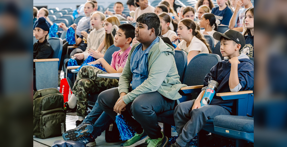 A group of people sitting in an auditorium, some holding blue pom-poms and backpacks.