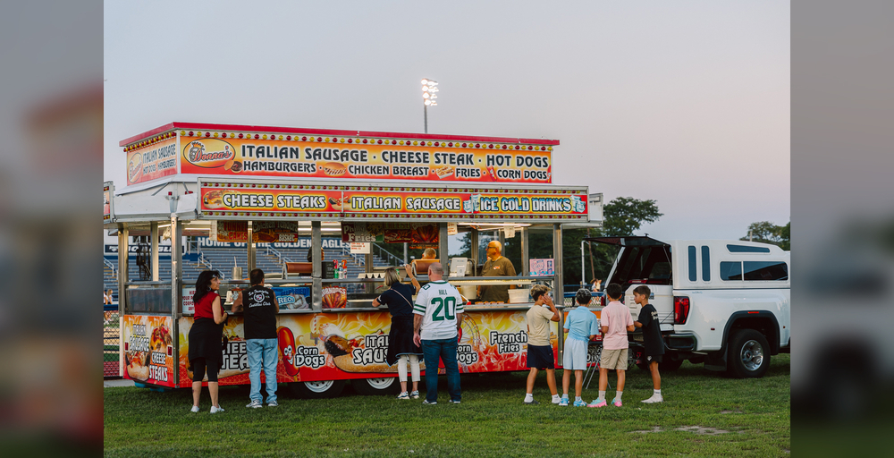 People gather at a food truck serving various items in a grassy area during an