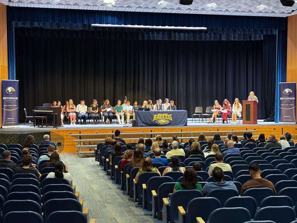 Students sitting on stage in auditorium with family members in audience.