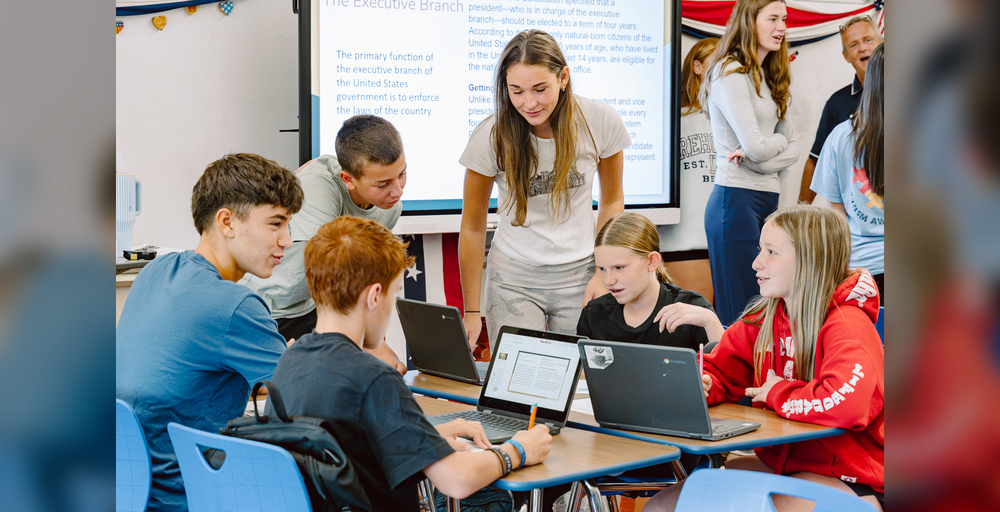 A group of students collaborate around laptops in a classroom setting with a presentation screen.
