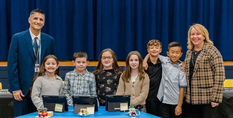 A group of six smiling children stands behind a blue table with tablets and colorful models. Two adults in business attire stand on either side, conveying a celebratory tone.