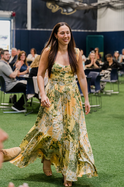 Young woman in a yellow floral dress walking gracefully at a lively indoor event, surrounded by seated guests.