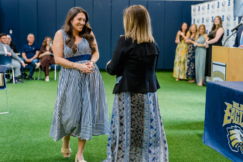 Two women celebrate a moment during a ceremony, with an audience in the background, highlighting a joyful award presentation on a green indoor field.