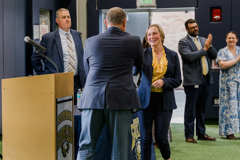 Event attendees celebrate as a woman shakes hands with a speaker at a podium, receiving recognition during a formal ceremony at an educational institution.