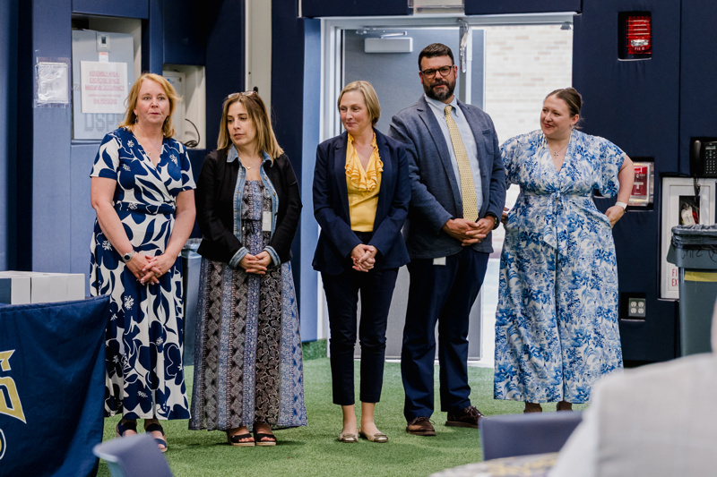 Group of five professionals standing together at an indoor event, dressed in various attire, showcasing a collaborative atmosphere.