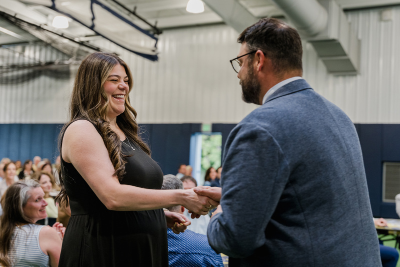 A woman in a black dress smiles while shaking hands with a man in a blue suit during a lively event, with an audience engaged in the background.