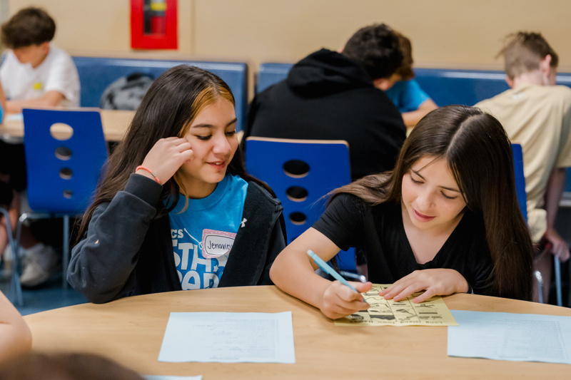 Two girls engage in a fun activity at a classroom table, one smiling while the other writes on a worksheet. The background shows other students participating in the same event.