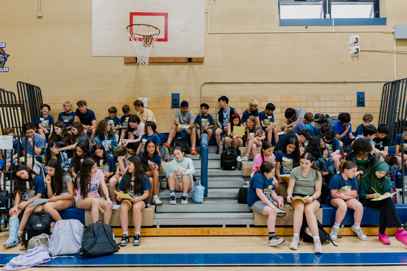 Students sitting on bleachers in a gymnasium, engaged in various activities, including reading and socializing, with a basketball hoop in the background.