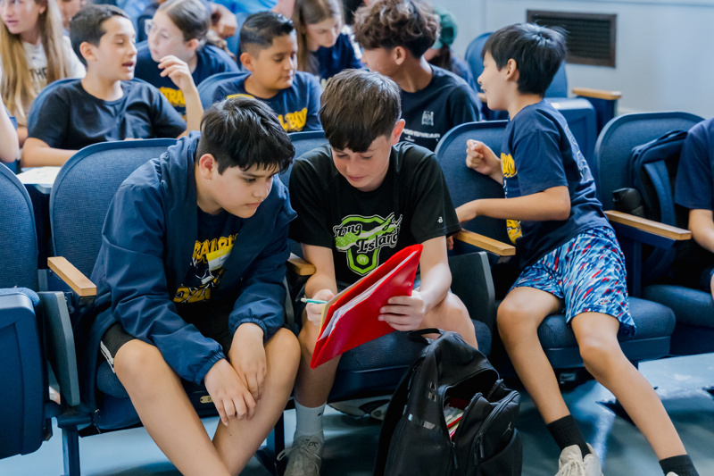 Two students collaborate while sitting in a classroom, surrounded by peers, with one holding a red folder and the other focused on a notebook.