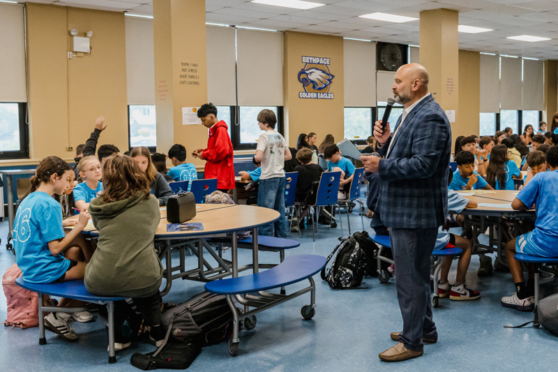 Principal addressing students in a busy school cafeteria, with groups of children engaging at tables and wearing blue shirts, showcasing a vibrant educational environment.