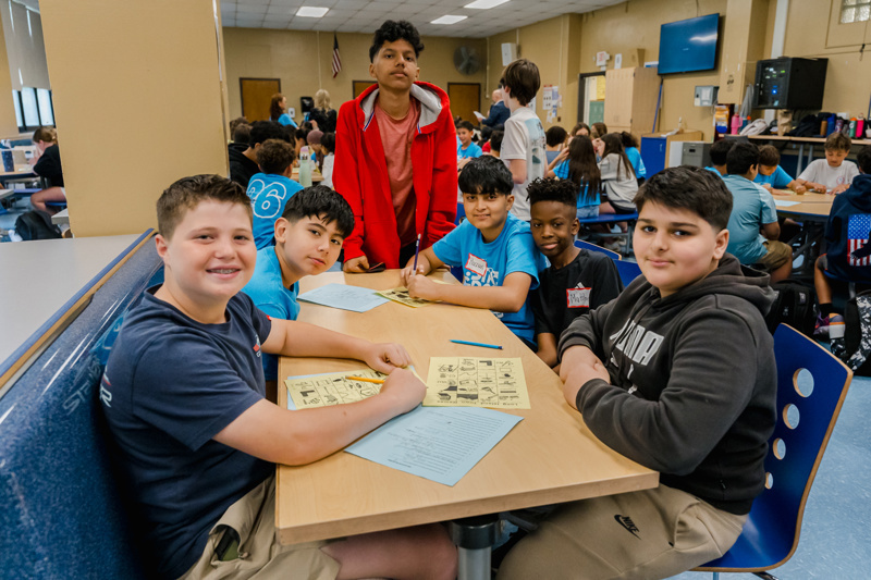 Students collaborate at a table in a lively classroom, engaging in a group activity with worksheets, fostering teamwork and learning in a vibrant educational environment.
