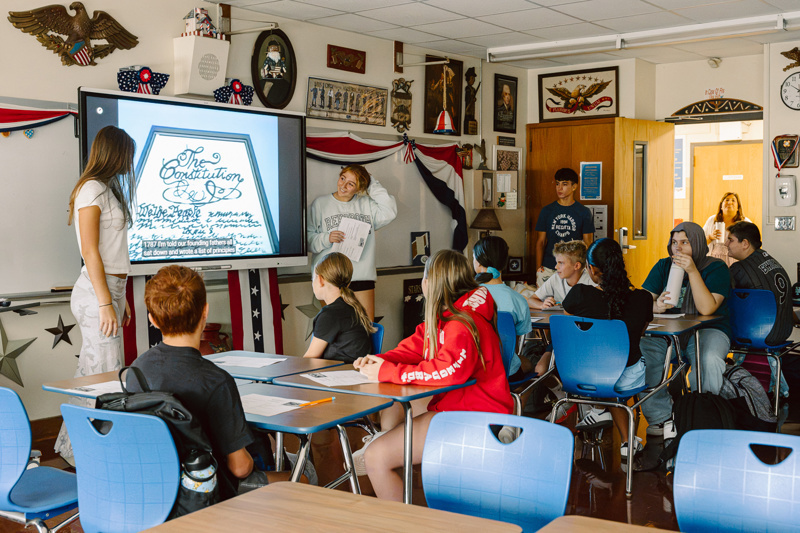 A group of people in a classroom watching a presentation on a screen displaying text.