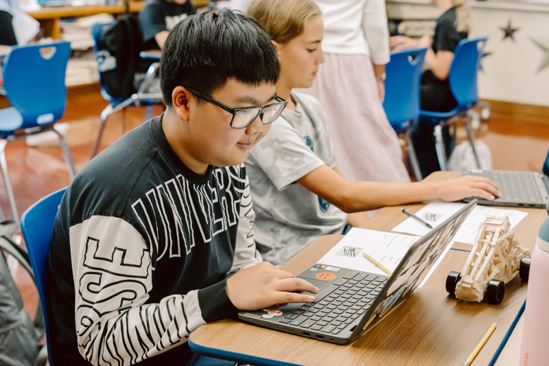 Students working on laptops at desks in a classroom with various objects and materials.
