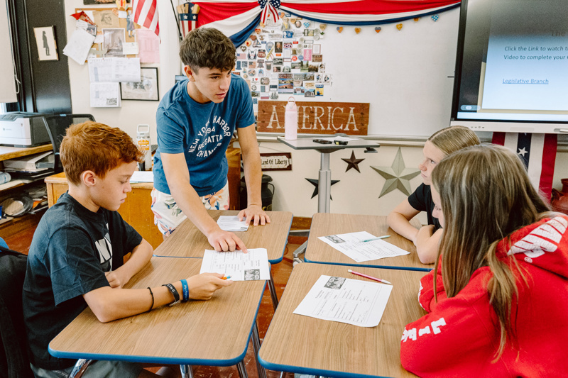 A group of students collaborate at a table, discussing papers in a classroom setting.