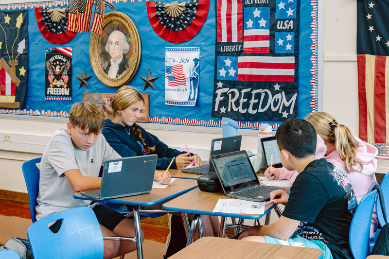 Students working at desks with laptops in a classroom decorated with patriotic posters.