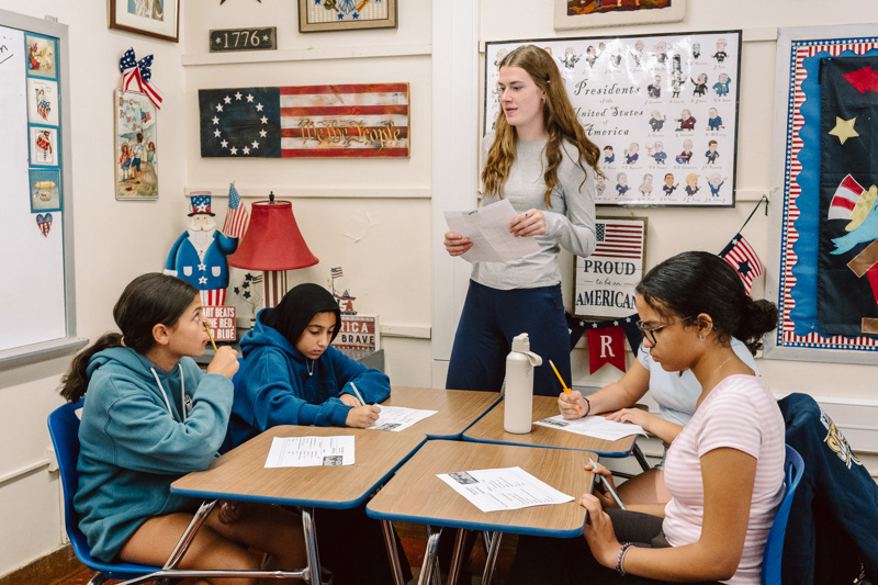A group of people sit at desks while another person stands, holding papers in a
