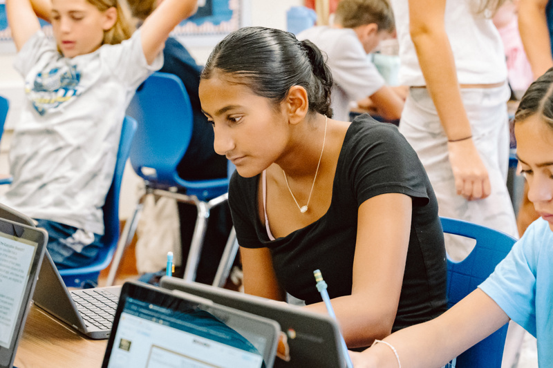 Students working on laptops in a classroom with blue chairs and classroom materials visible.