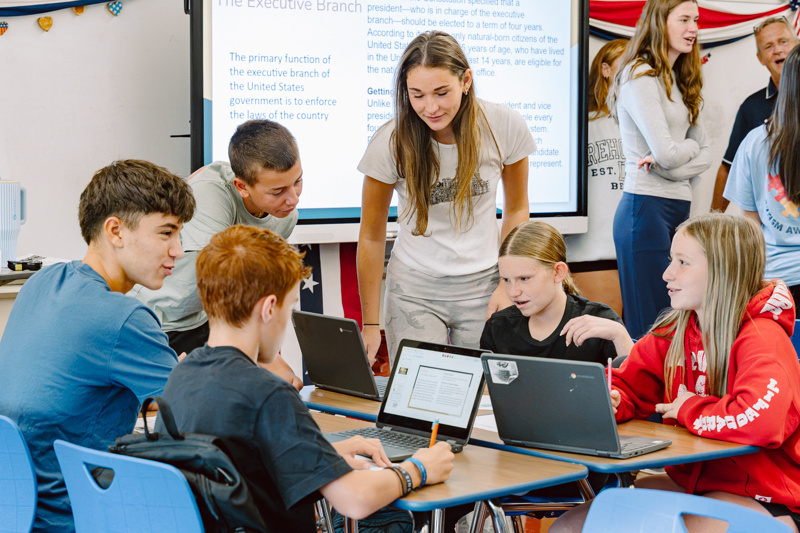 Students working together with laptops in a classroom, with a presentation displayed on a screen.