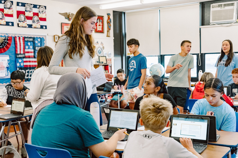 Group of people in a classroom using laptops and in conversation with a teacher.