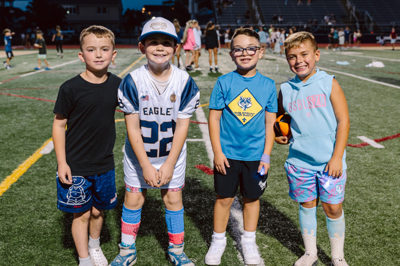 Four children pose on a sports field, wearing casual clothing and holding a ball.