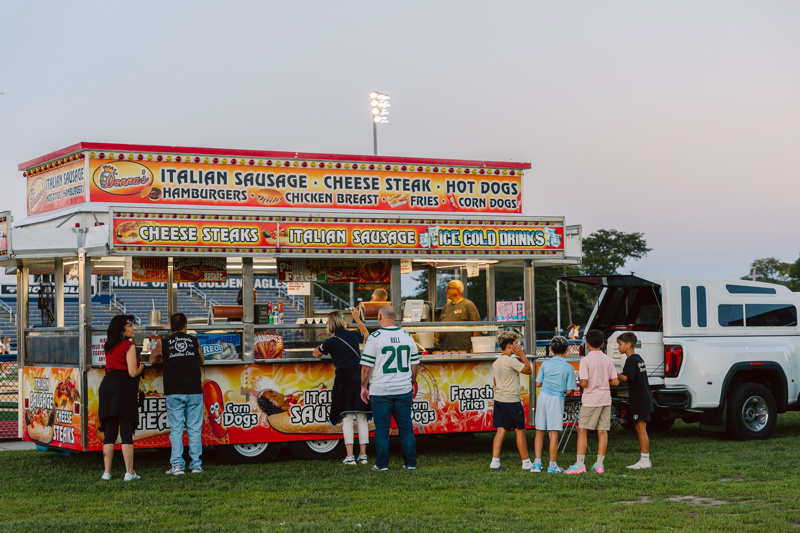 People gather around a colorful food truck serving various snacks and beverages in a field.