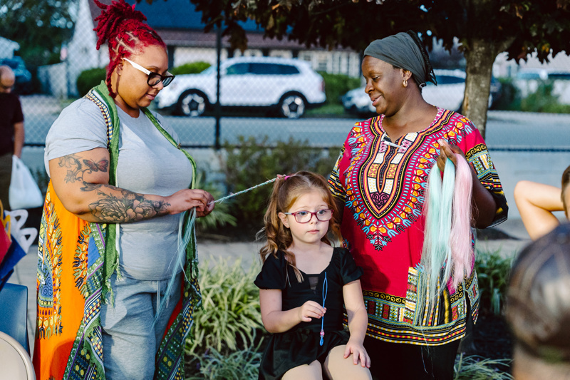 A group of people working with colored hair extensions outdoors, with vehicles in the background.