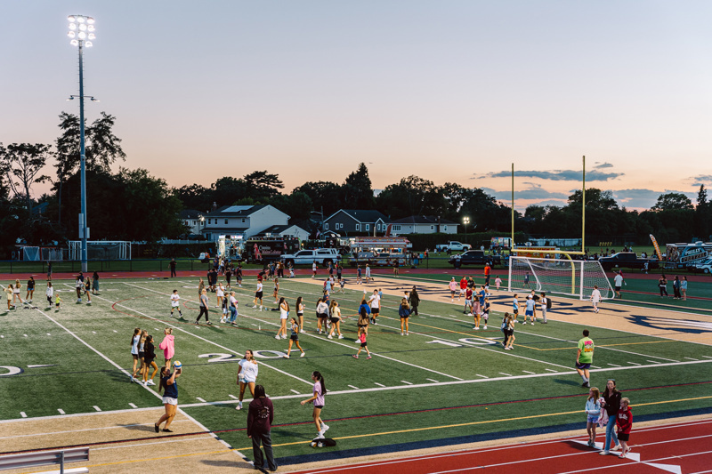 People on a sports field with goalposts, lights, and soccer equipment during dusk.