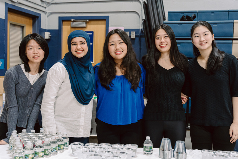 People standing behind a table with drinks and cups in a gymnasium setting.