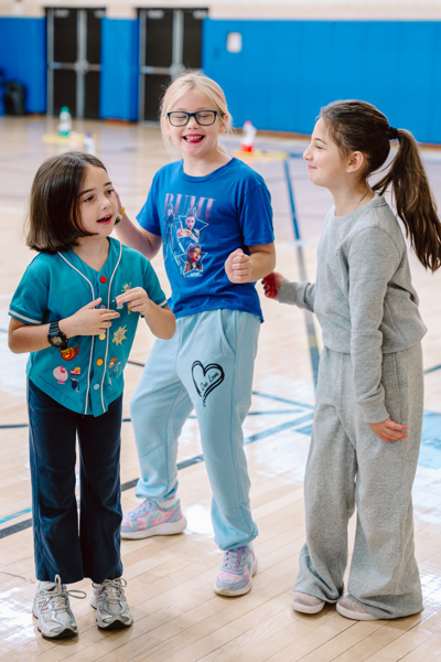 Three people stand together in a gym, wearing casual clothing and interacting with each other.