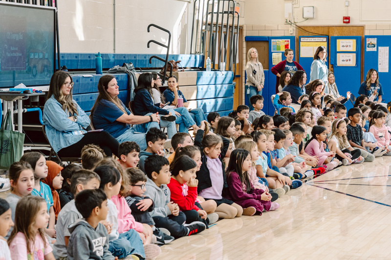 A group of children sitting on the floor while adults observe in a school gymnasium.