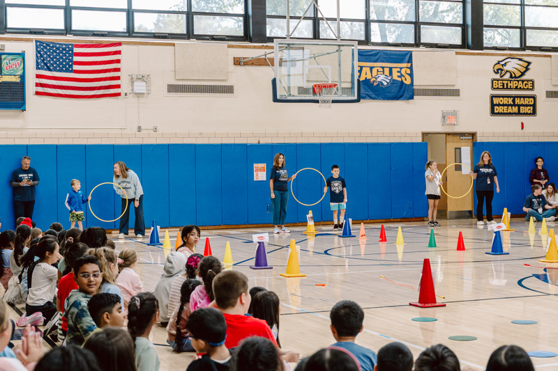 Children participate in gym activities with cones and hula hoops in a school gymnasium.