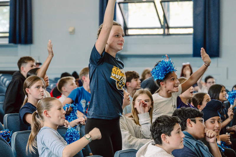 A group of people sitting in an auditorium, some holding blue pom-poms and backpacks.