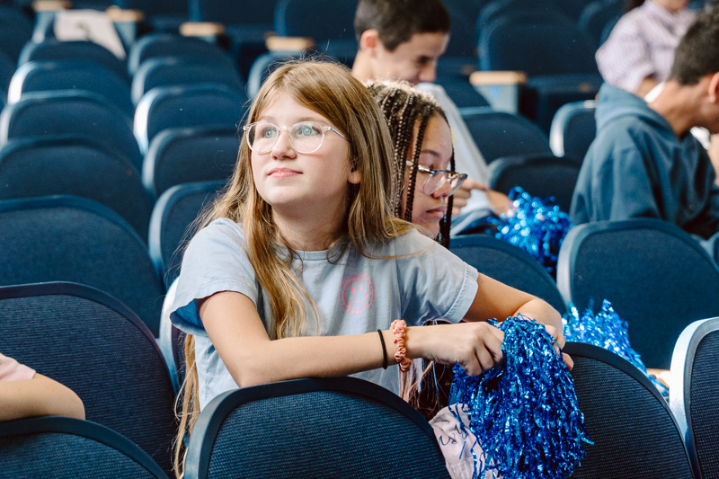 A group of people sitting in an auditorium, some holding blue pom-poms and backpacks.