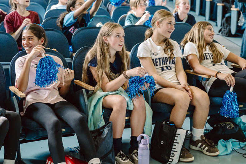 A group of people sitting in an auditorium, some holding blue pom-poms and backpacks.