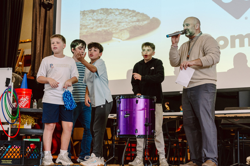 A group performs on stage, featuring a drum, props, and a large pizza backdrop.